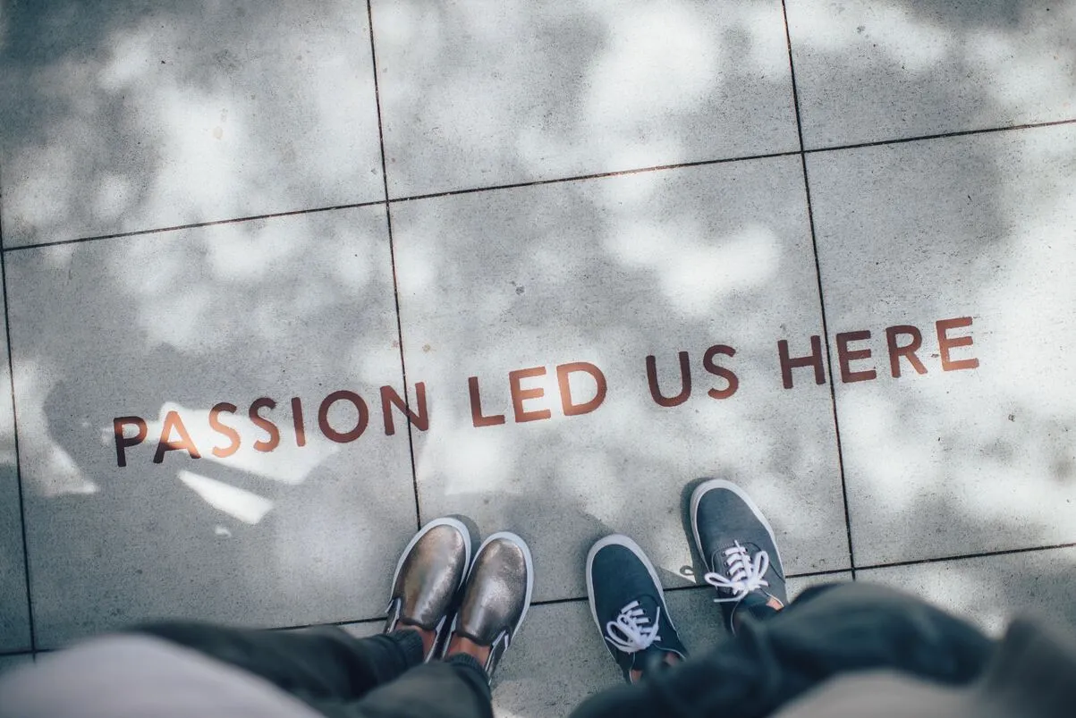 Photo Credit: Ian Schneider for Unsplash. Photo showing the feet of two people infront of the writing on large cement squares "Passion Led Us Here" with shadows of the overhead trees 